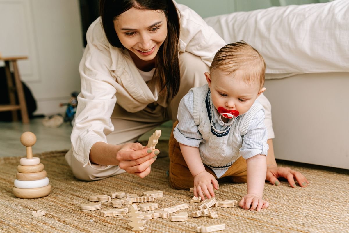 Baby crawling on a rug and playing with wooden toys while a parent sits beside him