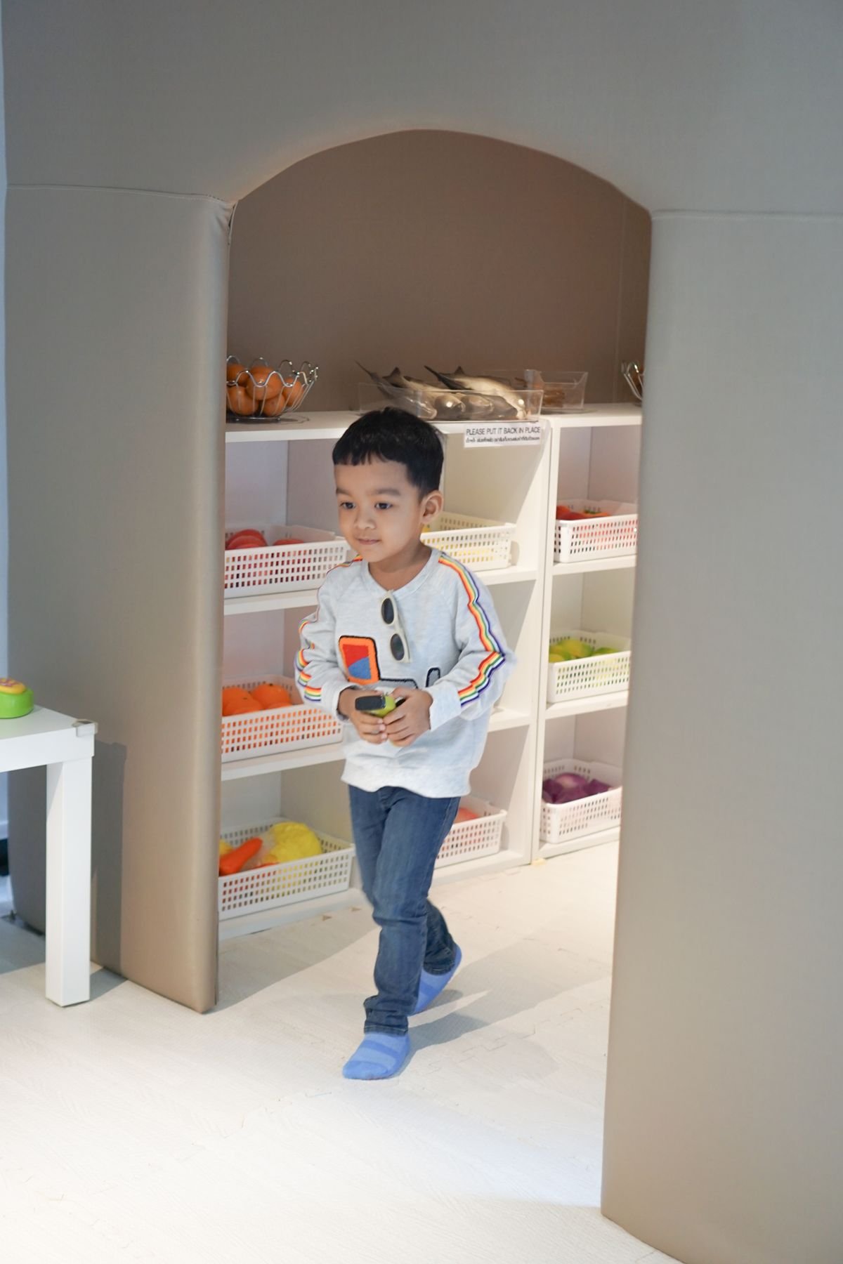Young boy carrying a toy past open shelves with labeled bins in a playroom nook